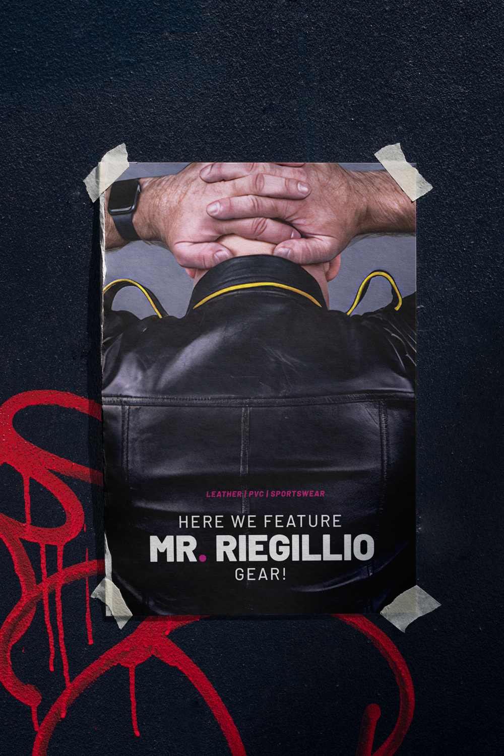 Poster featuring a person wearing a black leather jacket with hands clasped behind their head, taped to a wall with red graffiti.