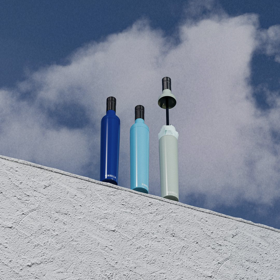 Three closed umbrellas in blue, light blue, and light gray colors standing upright on a textured white wall against a cloudy sky.