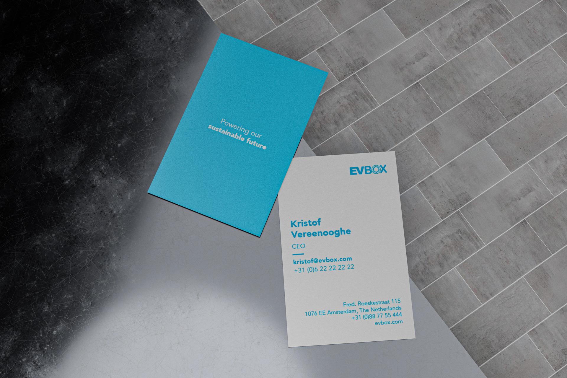 Two business cards on tiled floor; one blue card with 'Powering our sustainable future' and one white card with EVBox logo and contact details for Kristof Vereenooghe, CEO.