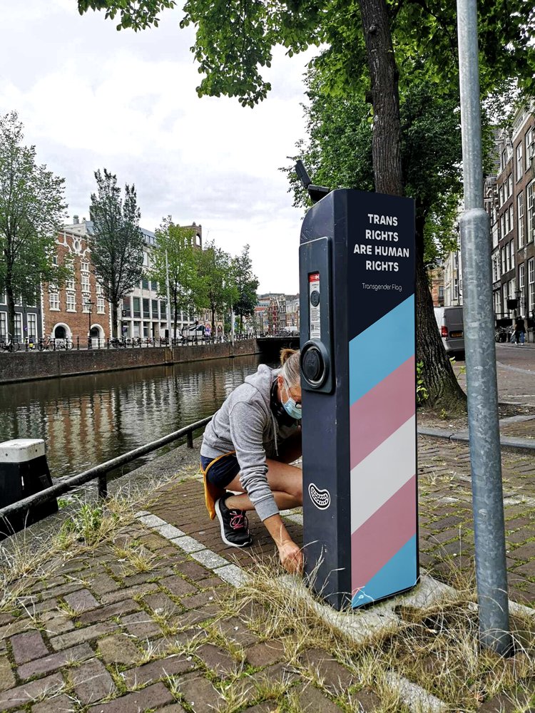 Person wearing a mask crouching by a canal next to a parking meter decorated with the transgender flag colors and the text 'Trans Rights Are Human Rights'.