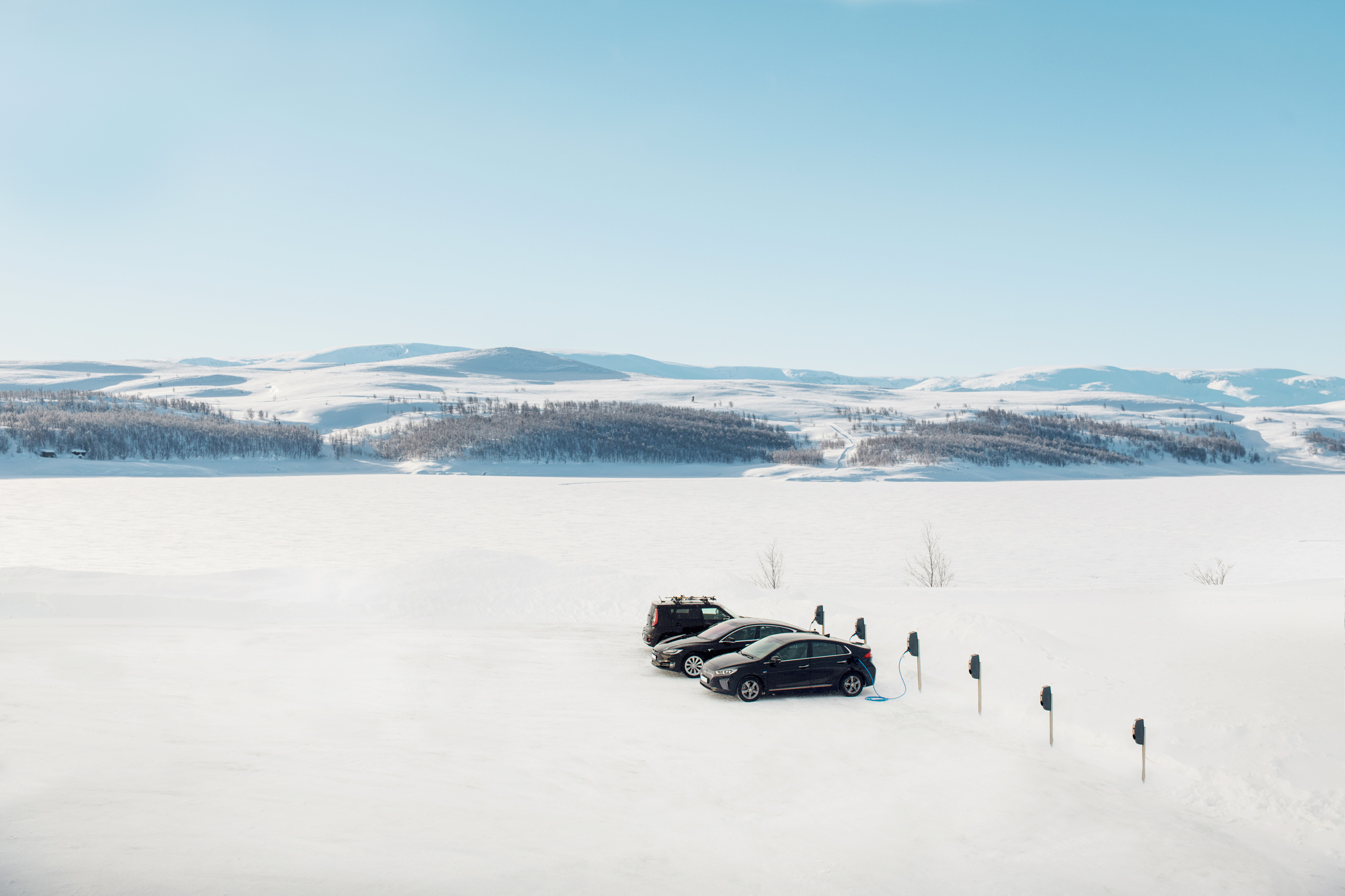 Two black electric cars parked and charging in a snowy landscape with snow-covered hills in the background under a clear blue sky.