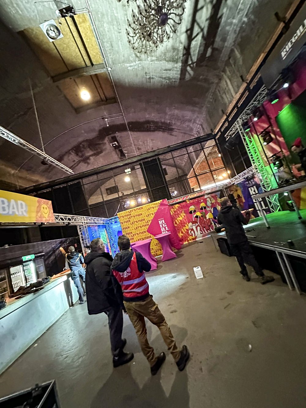 Interior of an event space with a high arched ceiling, colorful banners, stage lighting, and people standing near a bar and stage.