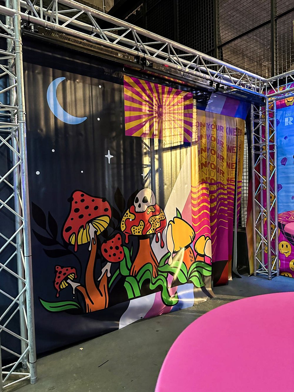 Colorful stage backdrop featuring whimsical mushrooms with one topped by a skull, a crescent moon, stars, and bold graphic patterns in yellow, pink, and purple hues.