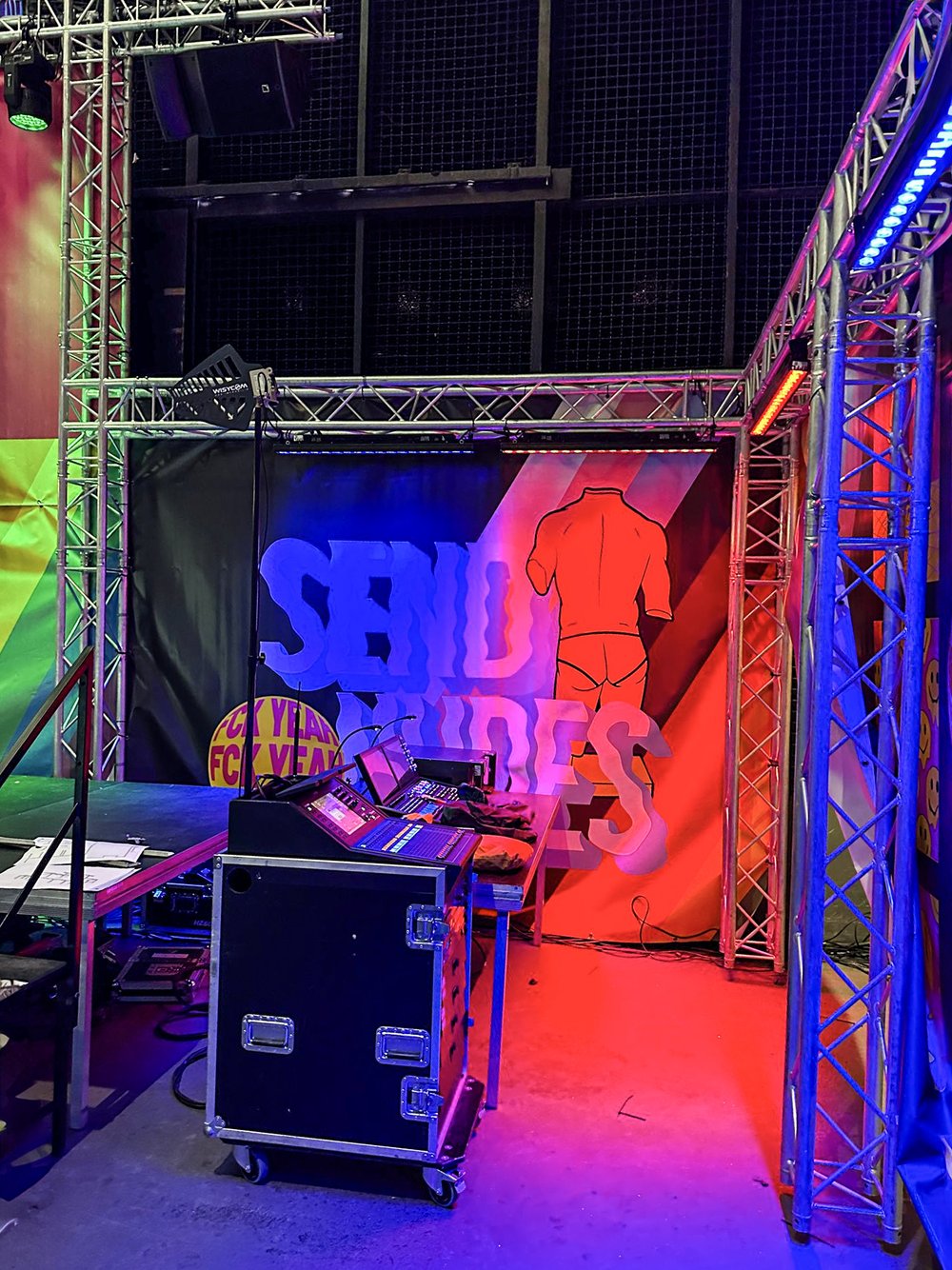  backstage area with sound mixing equipment, colorful stage lighting, and a backdrop featuring a headless red figure and large text partially reading 'SEND'.