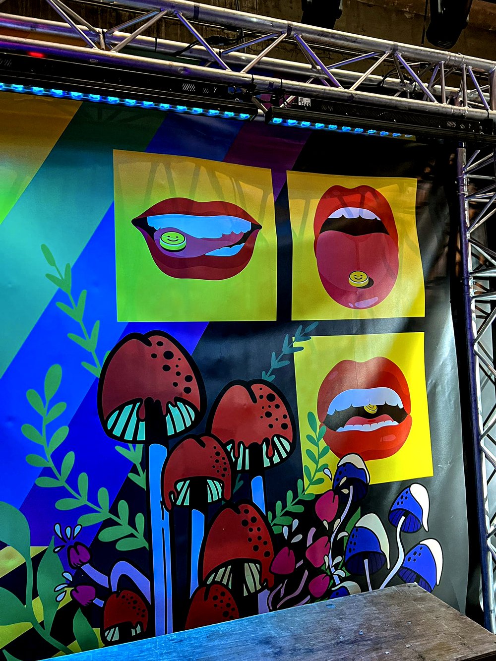 Colorful mural featuring red and blue mushrooms with green leaves and three yellow panels showing illustrations of lips holding smiley face pills.