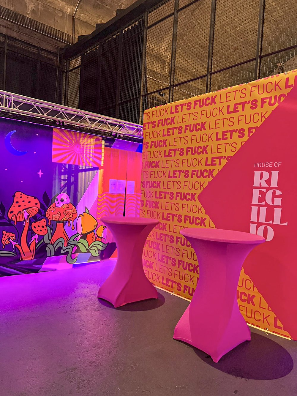 Two pink high cocktail tables in front of bright yellow and pink wall with repeating text 'LET'S FUCK' and a red geometric shape with 'HOUSE OF RIEGILLO' text, next to colorful mural featuring red mushrooms, a skull, and a crescent moon.