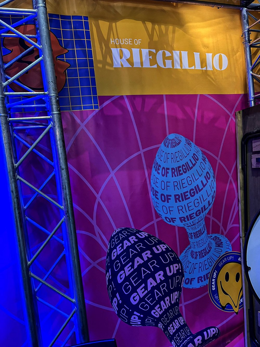 Stage design with metal scaffolding, a yellow banner reading 'HOUSE OF RIEGILLIO,' and two large abstract shapes covered in text: one blue with repeated 'HOUSE OF RIEGILLIO' and one dark with 'GEAR UP!' repeated.