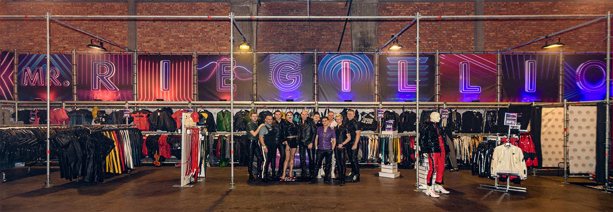Group of nine people in leather clothing standing in front of a clothing booth with neon signs spelling MR. RIEGELLO.