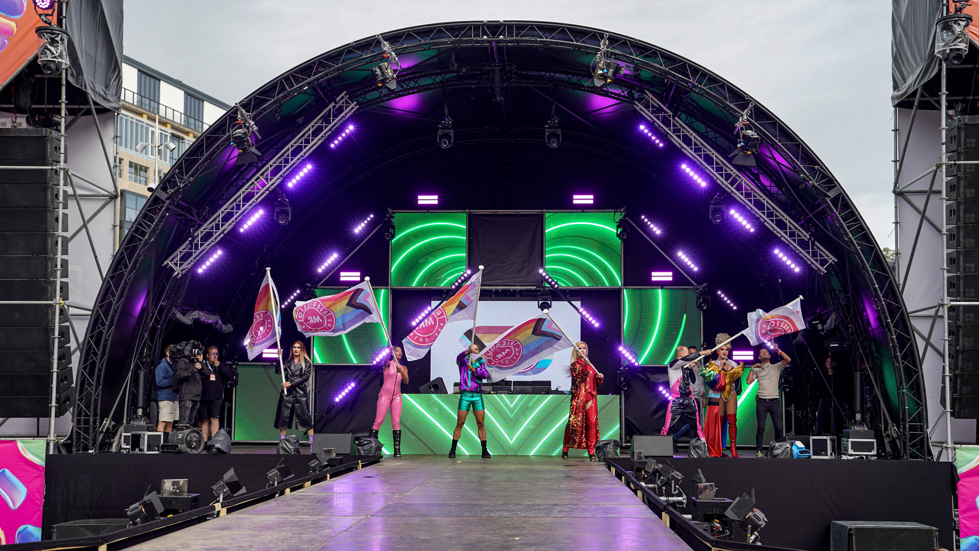 Six performers on stage under an arch of purple lights waving pride-themed flags, with a DJ setup and green neon heart visuals behind them.