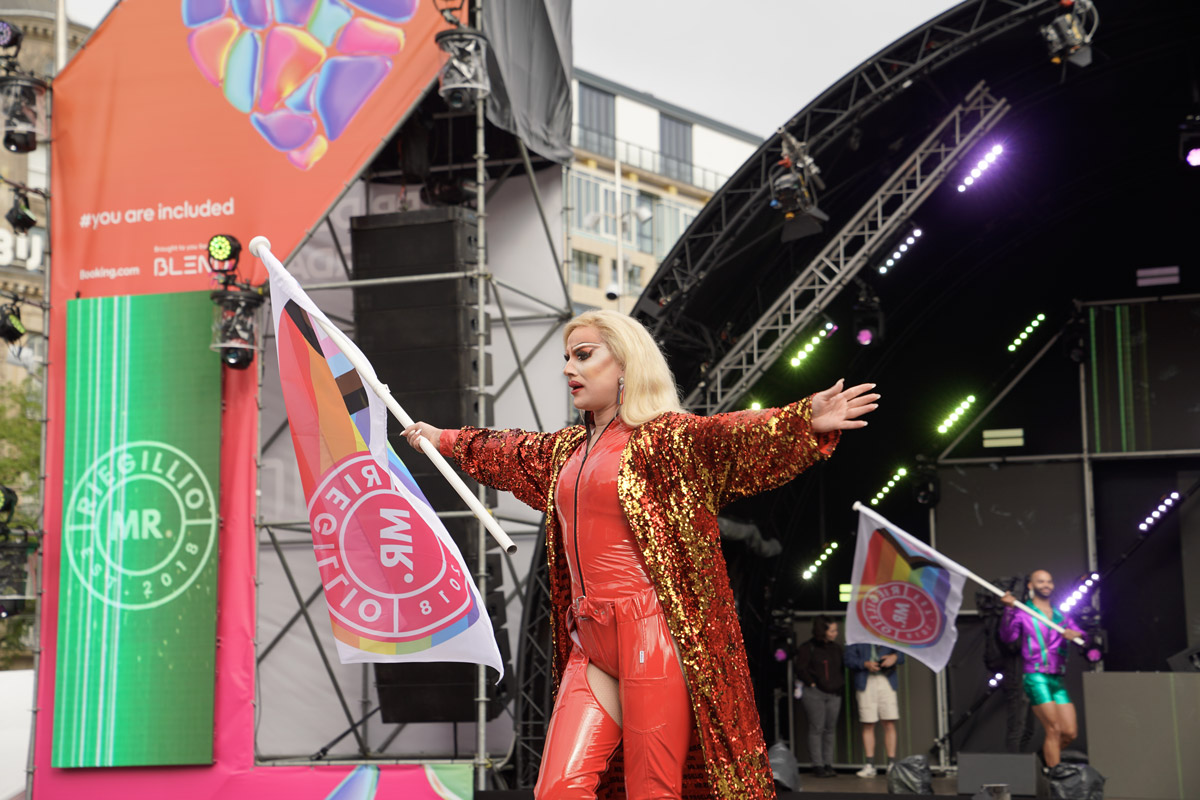 Person in shiny red outfit and sequined coat holding a flag on stage at a pride event.