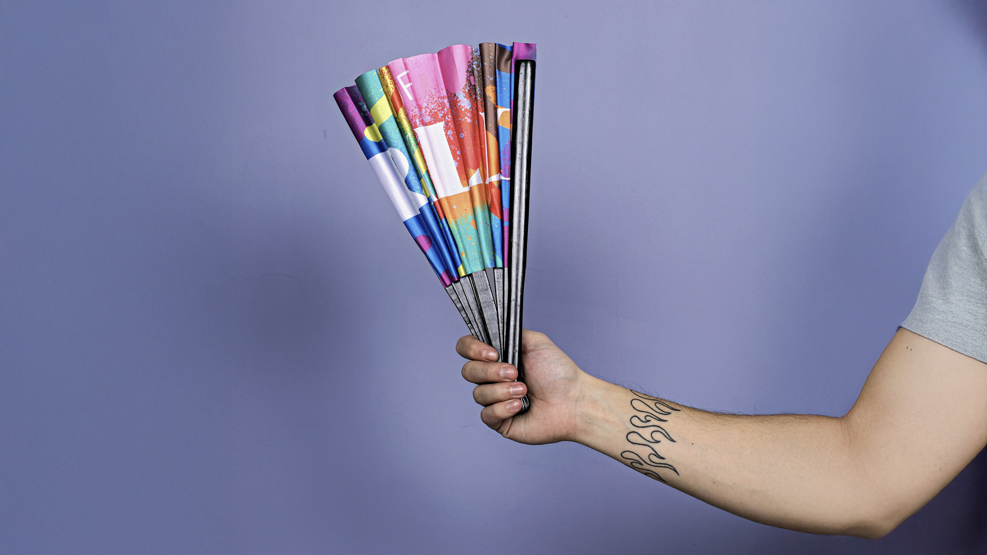 Person's arm with flame tattoo holding a closed colorful folding fan against a purple background.