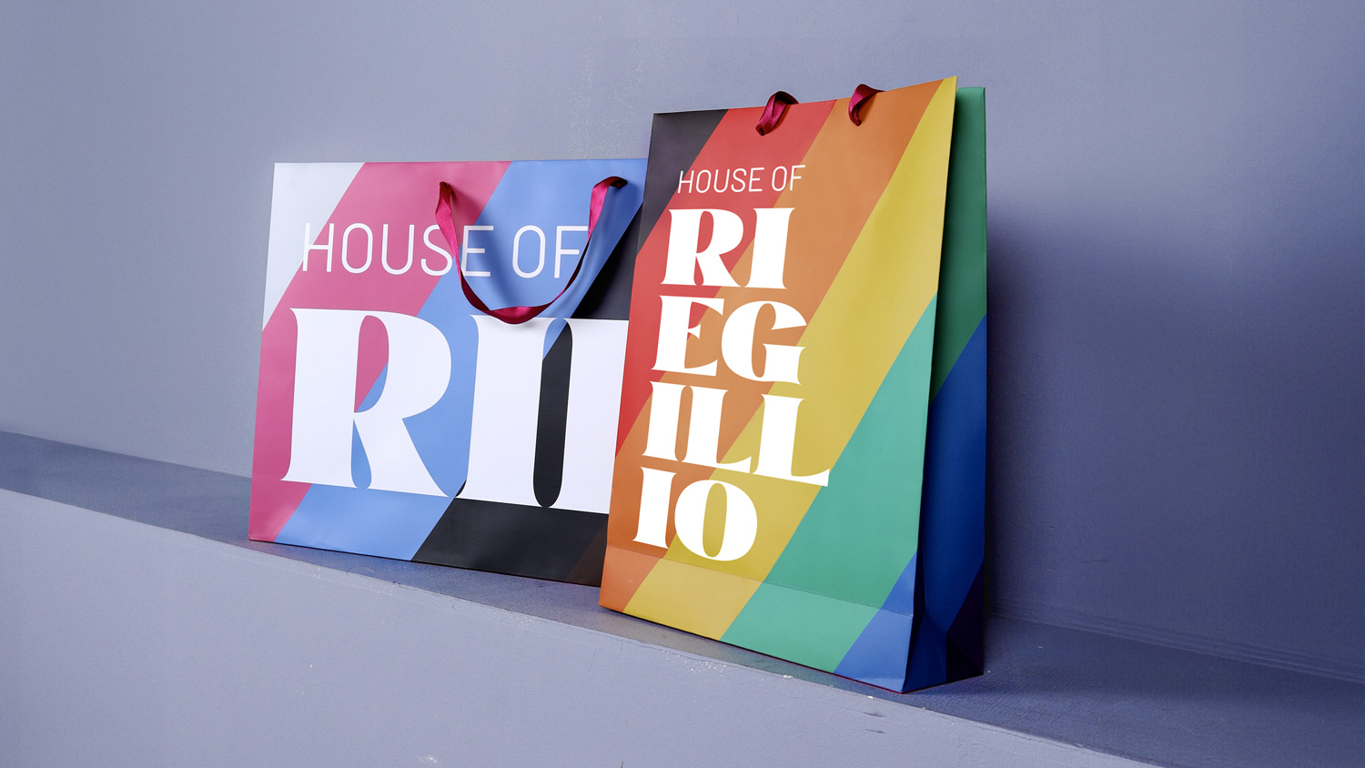 Two colorful shopping bags with diagonal stripes and the text 'HOUSE OF RIELLIO' on a smooth blue-gray surface against a matching background.
