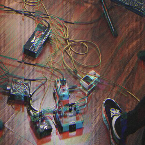 Multiple guitar effects pedals connected by cables on a wooden floor next to a person's foot wearing a black and white shoe.