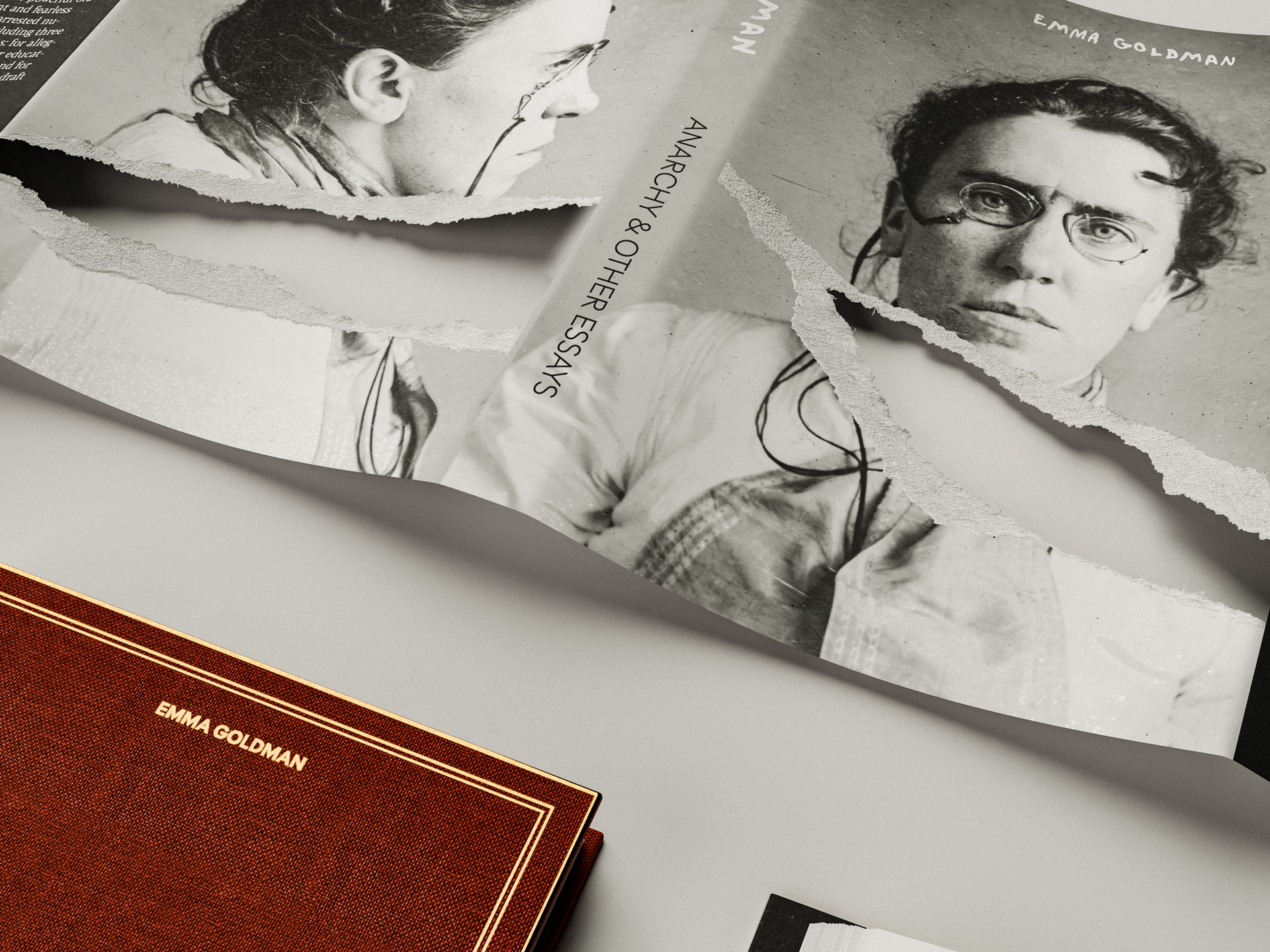 Torn black-and-white photograph of Emma Goldman with eyeglasses beside a red book titled 'Emma Goldman'.